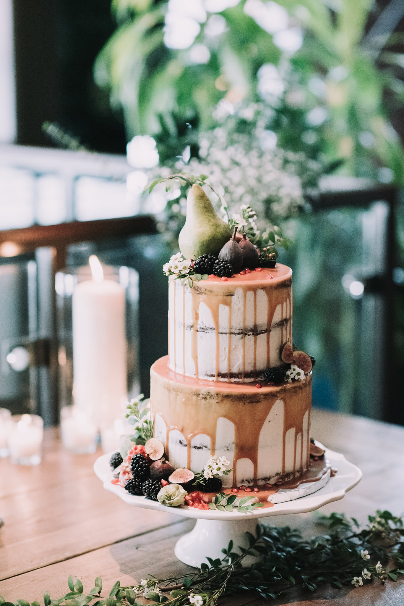 A rustic looking, two tier semi-naked cake with white cream cheese icing, caramel drip and fresh fruit adornments: pear, fig, blackberries and flowers